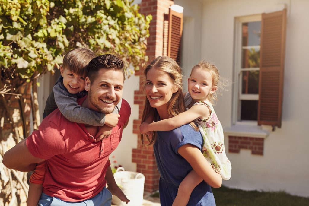 Happy father and mother looking at camera while piggybacking their son and daughter. Happy young family of four playing in their backyard on a sunny day.